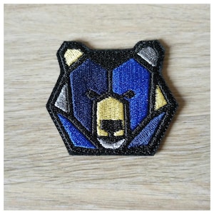 Może przedstawiać: Naszywka z haftowanym geometrycznym niedźwiedziem w kolorze niebieskim, żółtym i szarym. Pysk niedźwiedzia jest obwiedziony czarnym kolorem.