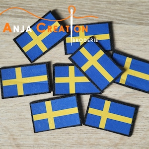 以下が含まれることがあります： 黒色のステッチが縁取りされた、黄色と青色のスウェーデン国旗のパッチが7つ集められています。パッチは木製の表面に積み重ねられています。画像の左上隅には「Anja Creation Broderie」というテキストが表示されています。