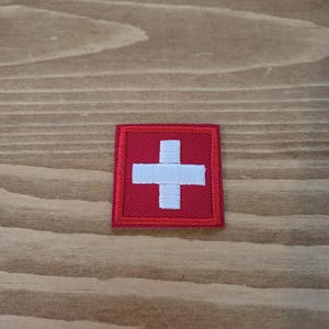 Op de afbeelding: Een vierkante, geborduurde patch met de Zwitserse vlag. De patch heeft een rode achtergrond met een wit kruis in het midden. Het kruis is gecentreerd in het rode vierkant, waarschijnlijk voor kleding of accessoires.
