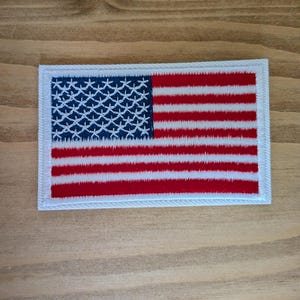 Peut inclure: Un écusson rectangulaire brodé représentant le drapeau des États-Unis. L'écusson présente un canton bleu foncé avec des motifs d'étoiles blanches brodées et des bandes horizontales rouges et blanches alternées, bordé d'un liseré blanc.