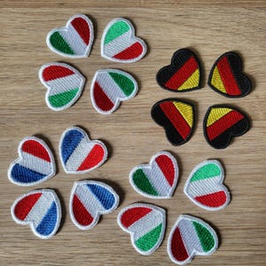May include: Embroidered heart-shaped patches featuring the flags of Italy, Germany, France, and Belgium. The patches have a white background with red, green, yellow, and blue accents. The patches are arranged on a wooden surface.