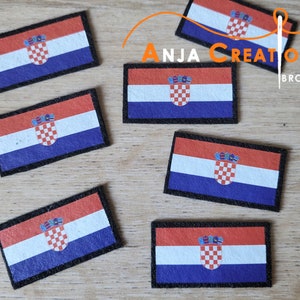 May include: Six Croatian flag patches with a black border. The patches are made of fabric and have a white background with a red and white checkered shield in the center. The shield is topped with a crown. The patches are arranged in a staggered pattern on a wooden surface.