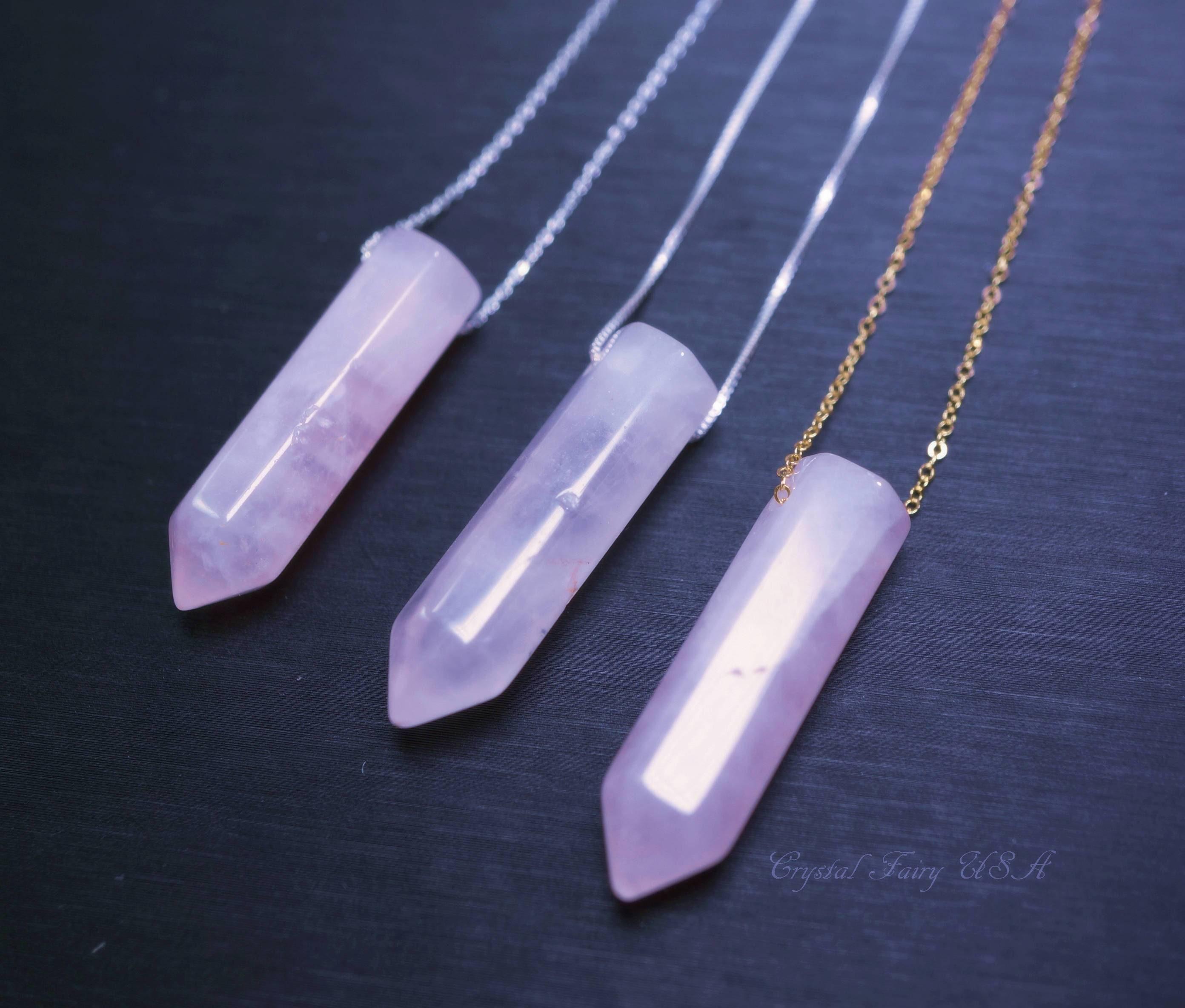 May include: Three rose quartz crystal pendant necklaces. Each pendant is a light pink, elongated hexagonal shape with a pointed tip. The necklaces have silver and gold chains. The background is a dark, solid color.