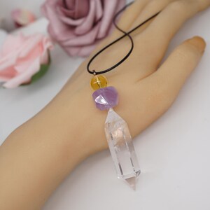 May include: A clear quartz crystal pendant necklace with a black cord. The necklace features a yellow bead, a purple faceted bead, and a clear quartz crystal point. The necklace is displayed on a light-toned surface.