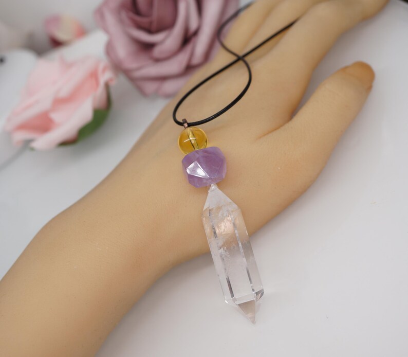 May include: A clear quartz crystal pendant necklace with a black cord. The necklace features a yellow bead, a purple faceted bead, and a clear quartz crystal point. The necklace is displayed on a light-toned surface.