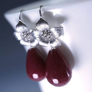 May include: Silver-tone flower earrings with teardrop-shaped, deep red gemstones. The earrings feature a floral design at the top with a hook closure. The gemstones are a rich, dark red color, adding a touch of elegance.