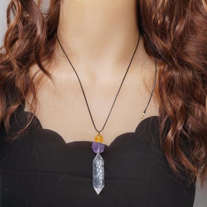 May include: A black cord necklace featuring a clear crystal pendant with a faceted design. The necklace also includes a purple and yellow gemstone accent. The pendant is suspended from a black cord.