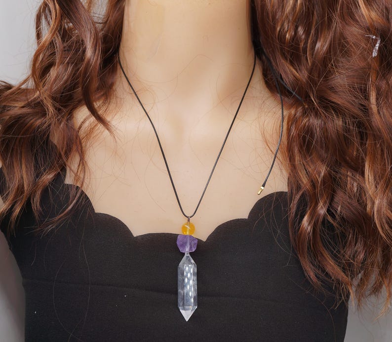 May include: A black cord necklace featuring a clear crystal pendant with a faceted design. The necklace also includes a purple and yellow gemstone accent. The pendant is suspended from a black cord.