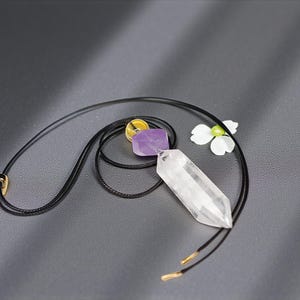 May include: A black cord necklace featuring a clear quartz crystal pendant, a purple amethyst stone, and a yellow and gold accent. A white flower is placed near the necklace on a gray surface. The necklace has gold-colored metal accents.