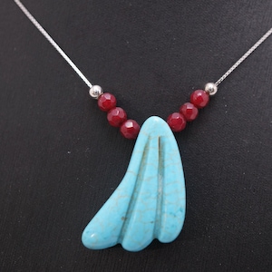May include: A silver necklace featuring a turquoise pendant with a carved design. The pendant is accented with red beads and a silver chain. The necklace is displayed against a black background.