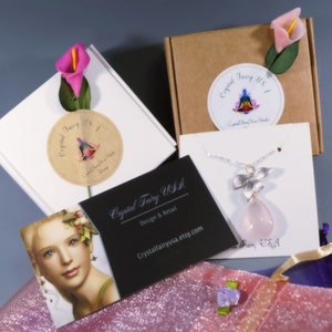 May include: An overhead shot of jewelry packaging and a necklace. The necklace features a silver flower pendant with a pink teardrop-shaped stone. The packaging includes white and brown boxes, business cards, and a pink glittery bag. The text "Crystal Fairy USA" is visible.