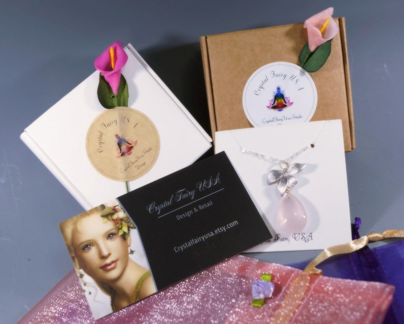 May include: An overhead shot of jewelry packaging and a necklace. The necklace features a silver flower pendant with a pink teardrop-shaped stone. The packaging includes white and brown boxes, business cards, and a pink glittery bag. The text "Crystal Fairy USA" is visible.