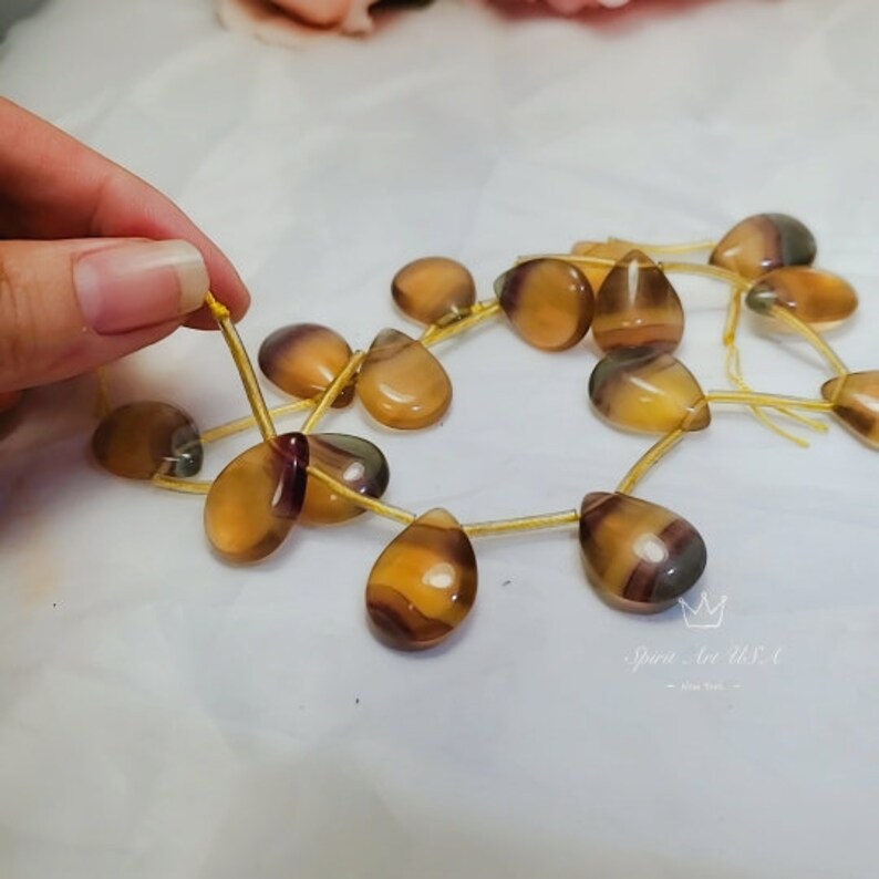 以下が含まれることがあります： 茶色、黄色、紫色の混合色をした涙滴型のビーズの連なり。ビーズは金色の紐に通され、円形に配置されています。