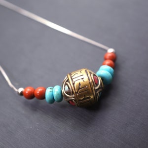 May include: A silver chain necklace with a large, round, gold bead with engraved lettering. The bead is surrounded by smaller turquoise and red beads.