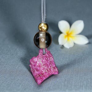 May include: A handmade necklace featuring a raw, textured, pink gemstone pendant. The pendant is suspended from a gray cord, with a gold-colored bead and a dark brown and clear bead. A white flower with a yellow center is in the background.