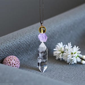 May include: A clear quartz crystal pendant necklace with a black cord. The pendant features a clear quartz crystal point, a purple amethyst, and a yellow bead. A small pink decorative sphere and white flowers are also in the image.