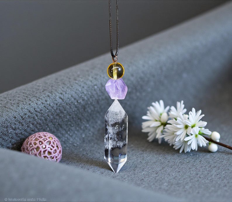 May include: A clear quartz crystal pendant necklace with a black cord. The pendant features a clear quartz crystal point, a purple amethyst, and a yellow bead. A small pink decorative sphere and white flowers are also in the image.