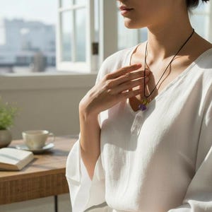 May include: A white linen top with a V-neck and long sleeves. A black cord necklace with a pendant featuring purple and yellow stones. A wooden table with an open book, a cup, and a small plant in a ceramic pot.
