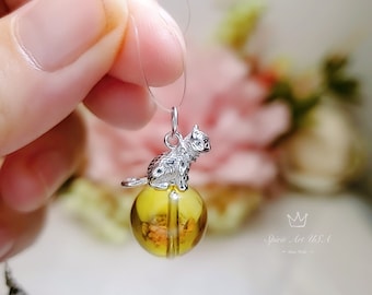 Peut inclure: Un pendentif rond en ambre jaune avec un charme de chat argenté. Le pendentif est attaché à une ficelle transparente.