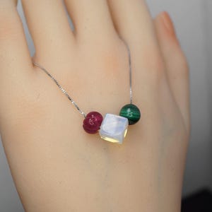 May include: A delicate silver necklace featuring a cube-shaped, iridescent gemstone, a carved red bead, and a green malachite bead. The necklace is displayed against a neutral background, highlighting the colors and textures of the gemstones.