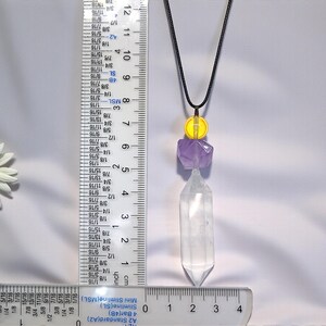 May include: A pendant necklace featuring a clear quartz crystal point, amethyst, and yellow bead accents. The necklace hangs from a black cord. The quartz crystal measures approximately 1.5 inches in length. The necklace is displayed against a white background.