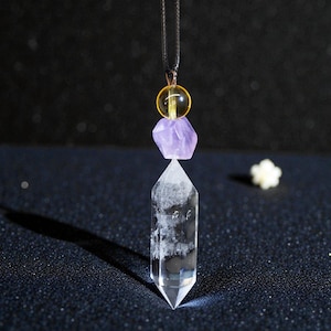 May include: A clear quartz crystal pendant with a faceted point, suspended from a black cord. The pendant features a purple amethyst and a yellow bead. The background is dark with a small white flower.