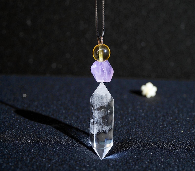 May include: A clear quartz crystal pendant with a faceted point, suspended from a black cord. The pendant features a purple amethyst and a yellow bead. The background is dark with a small white flower.