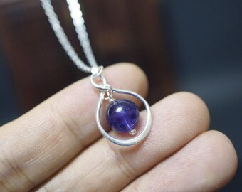 May include: A silver pendant necklace featuring a teardrop-shaped frame holding a deep purple, spherical gemstone. The necklace has a delicate silver chain. The gemstone is a rich, dark purple color, and the pendant is held in a hand.