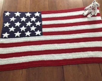 May include: A crocheted American flag blanket with red and white stripes and a blue square with white stars. A small stuffed animal with black spots rests on the blanket. The blanket is displayed on a wooden surface.