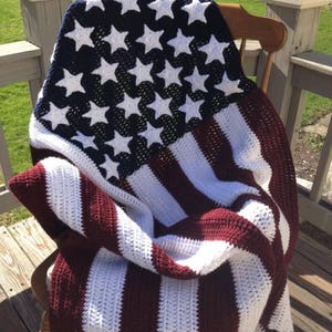 May include: A crocheted blanket draped over a wooden chair, featuring the design of the American flag. The blanket has a navy blue field with white stars and red and white stripes. The blanket is made of soft yarn.