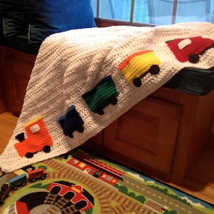 May include: A white crocheted blanket with a train theme. The blanket features five colorful train cars, each with a different color and design. The train cars are attached to the blanket with a white crocheted string.