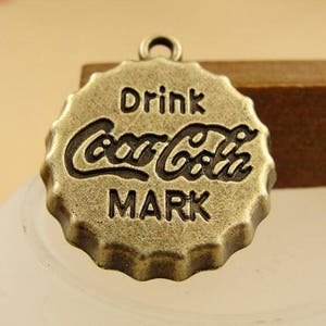 Może przedstawiać: Brązowy metalowy charm w kształcie zakrętki butelki z wygrawerowanym napisem "Drink Coca-Cola Mark".