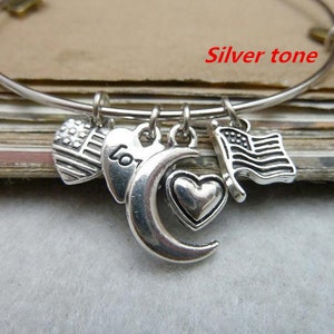 May include: Silver tone charm bracelet with a crescent moon, heart, and two American flag charms. The bracelet is on a brown surface.