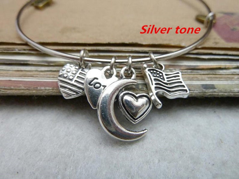 May include: Silver tone charm bracelet with a crescent moon, heart, and two American flag charms. The bracelet is on a brown surface.