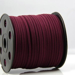 May include: A spool of burgundy suede cord. The cord is tightly wound around a black plastic spool. The suede cord is a deep, rich shade of burgundy, and the texture appears soft and smooth. The spool is set against a white background.