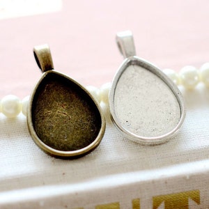 Könnte beinhalten: Zwei tropfenförmige Anhänger-Tabletts, eines silberfarben und eines bronzefarben, mit einer glatten, glänzenden Oberfläche. Die Tabletts sind so konzipiert, dass sie ein kleines Stück Glas oder anderes Material aufnehmen können.