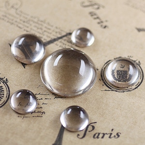 May include: Five clear glass cabochons of varying sizes on a brown paper background with the word "Paris" written in black ink.