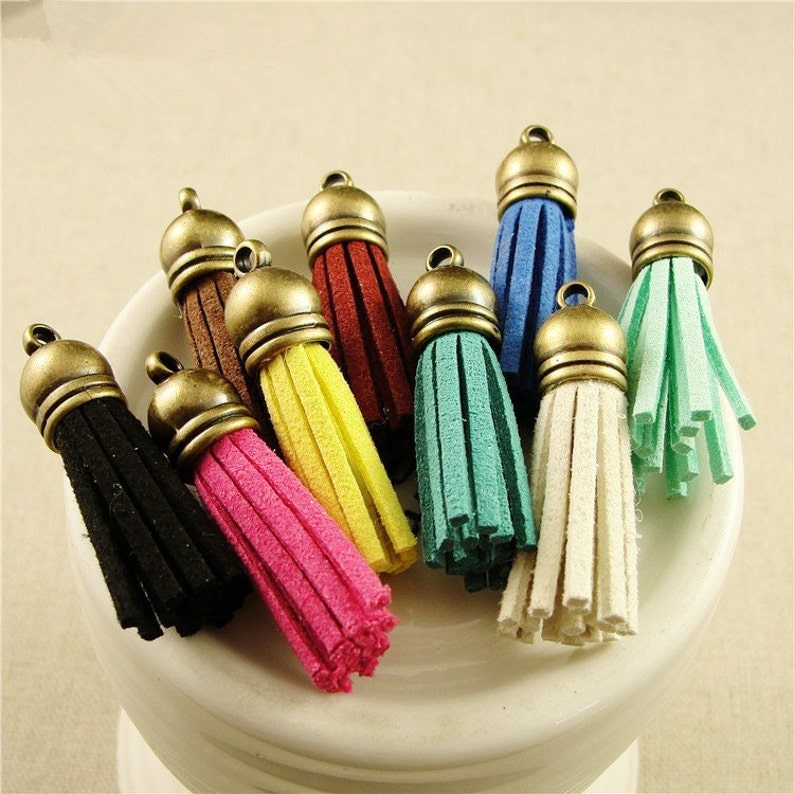 Puede incluir: Una colecci&oacute;n de borlas de gamuza de varios colores, cada una con una tapa de color bronce antiguo. Las borlas, en negro, marr&oacute;n, rosa, amarillo, rojo, turquesa, azul, blanco y verde, est&aacute;n dispuestas en un cuenco de cer&aacute;mica blanco. Adecuadas para la creaci&oacute;n de joyas y proyectos de artesan&iacute;a.