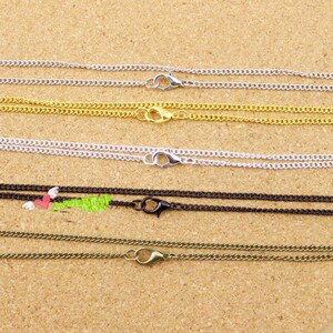 May include: Close-up of several necklace chains in various colors, including silver, gold, black, and bronze. Each chain has a lobster clasp for secure closure. The chains are displayed on a light brown surface.