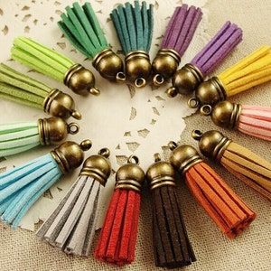Puede incluir: Una colecci&oacute;n de borlas de gamuza de varios colores, incluyendo verde, azul, morado y rojo. Cada borla tiene una tapa de color bronce con un peque&ntilde;o lazo para sujetar a joyas u otras manualidades. Las borlas est&aacute;n dispuestas en un patr&oacute;n circular.