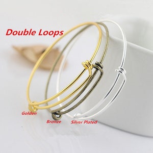May include: Three bangle bracelets in gold, bronze, and silver plated finishes. The bracelets are made of wire and have a double loop closure. The text "Double Loops" is visible at the top of the image.