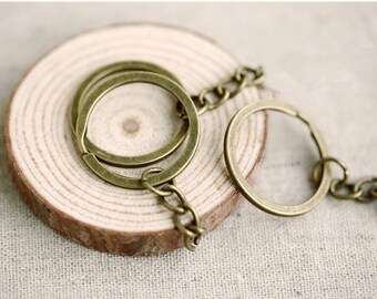 Könnte beinhalten: Ein Schlüsselanhänger aus Messing mit einem großen, runden Ring und einem kleineren, runden Ring, der an einer Kette befestigt ist. Der Schlüsselanhänger liegt auf einer Holzoberfläche.