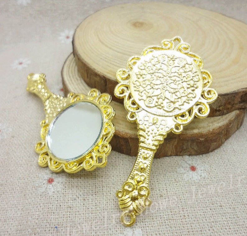 K&ouml;nnte beinhalten: Zwei verzierte Handspiegel in Goldton mit aufwendigen Designs. Ein Spiegel ist rund mit einer klaren reflektierenden Oberfl&auml;che, w&auml;hrend der andere ein aufwendigeres Design mit einem floralen Muster hat.