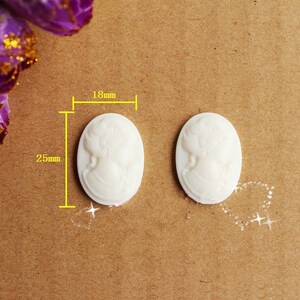 Puede incluir: Dos adornos de resina de estilo camafeo blancos, cada uno mide 18 mm de ancho y 25 mm de alto. Los adornos presentan un diseño en relieve de un perfil de mujer.