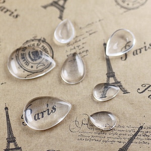 Könnte beinhalten: Sortiment aus klaren, tropfenförmigen Glascabochons, einige mit leichtem Schimmer, auf braunem Papieruntergrund mit einem gedruckten Design des Eiffelturms und dem Wort "Paris".