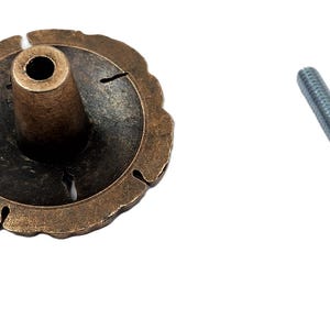 May include: A bronze-colored cabinet knob with a decorative, scalloped edge and a central post. A silver-colored screw is positioned to the right of the knob. The knob has a dark, aged appearance.