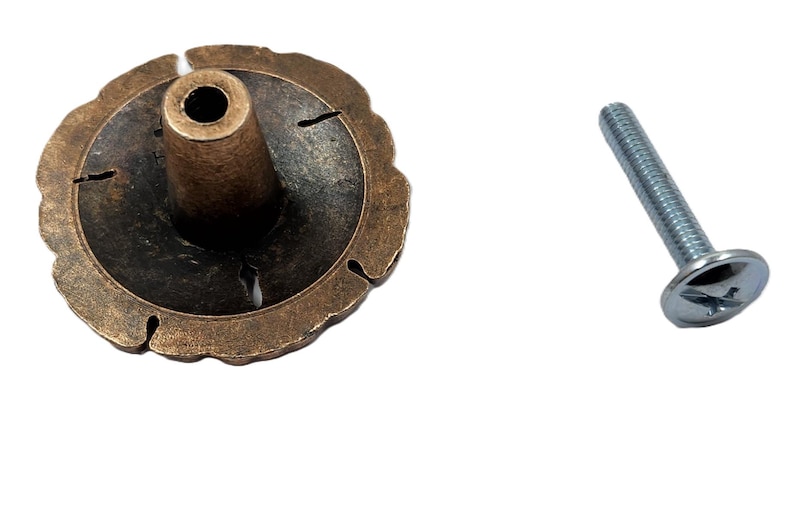 May include: A bronze-colored cabinet knob with a decorative, scalloped edge and a central post. A silver-colored screw is positioned to the right of the knob. The knob has a dark, aged appearance.