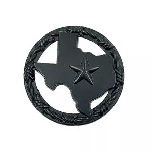 May include: A circular black metal emblem featuring a silhouette of the state of Texas with a centered five-pointed star. The entire design is enclosed within a textured border that mimics the appearance of twisted barbed wire fencing.