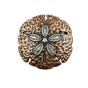 May include: A bronze-colored sand dollar with a blue and gold flower design. The sand dollar is textured and has a rustic look.