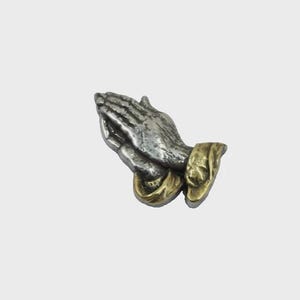 May include: A silver-toned metal sculpture of hands clasped in prayer, with gold-colored accents at the wrists. The hands are detailed with visible texture and shading, suggesting a vintage or antique aesthetic. The sculpture is set against a plain background.